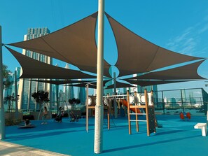 An outdoor play area with climbing structures, shaded by large canopies, and surrounded by a fence.