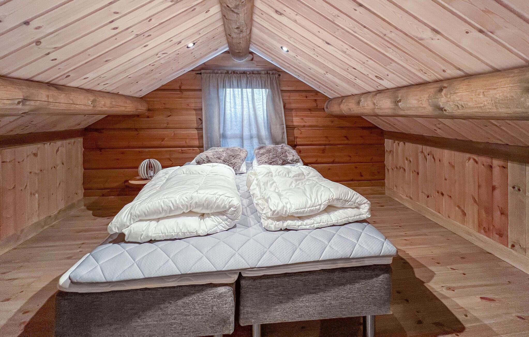 A cozy attic bedroom with a wooden sloped ceiling, a bed with two pillows, and a small bedside table.