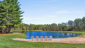 A serene lakeside setting with a row of beach chairs facing a calm pond, surrounded by lush greenery and tall trees.