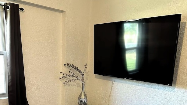 Une pièce dotée d’un téléviseur à écran plat fixé au mur, d’un meuble en bois foncé avec des tiroirs, d’un vase orné de branches et d’une fenêtre avec des rideaux noirs.