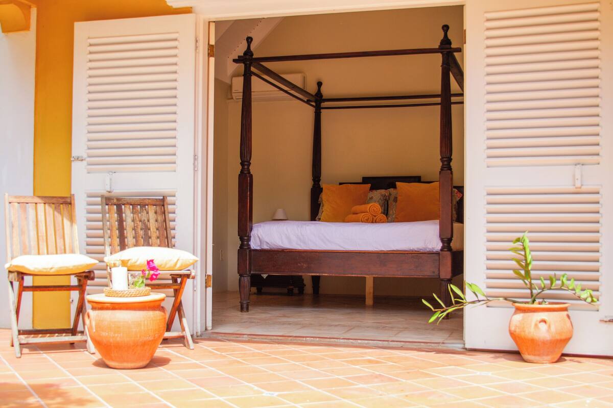Una cama con dosel con una colcha blanca y almohadas naranjas, una silla de madera con cojín, y dos macetas de terracota con plantas.
