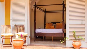 Una cama con dosel con una colcha blanca y almohadas naranjas, una silla de madera con cojín, y dos macetas de terracota con plantas.