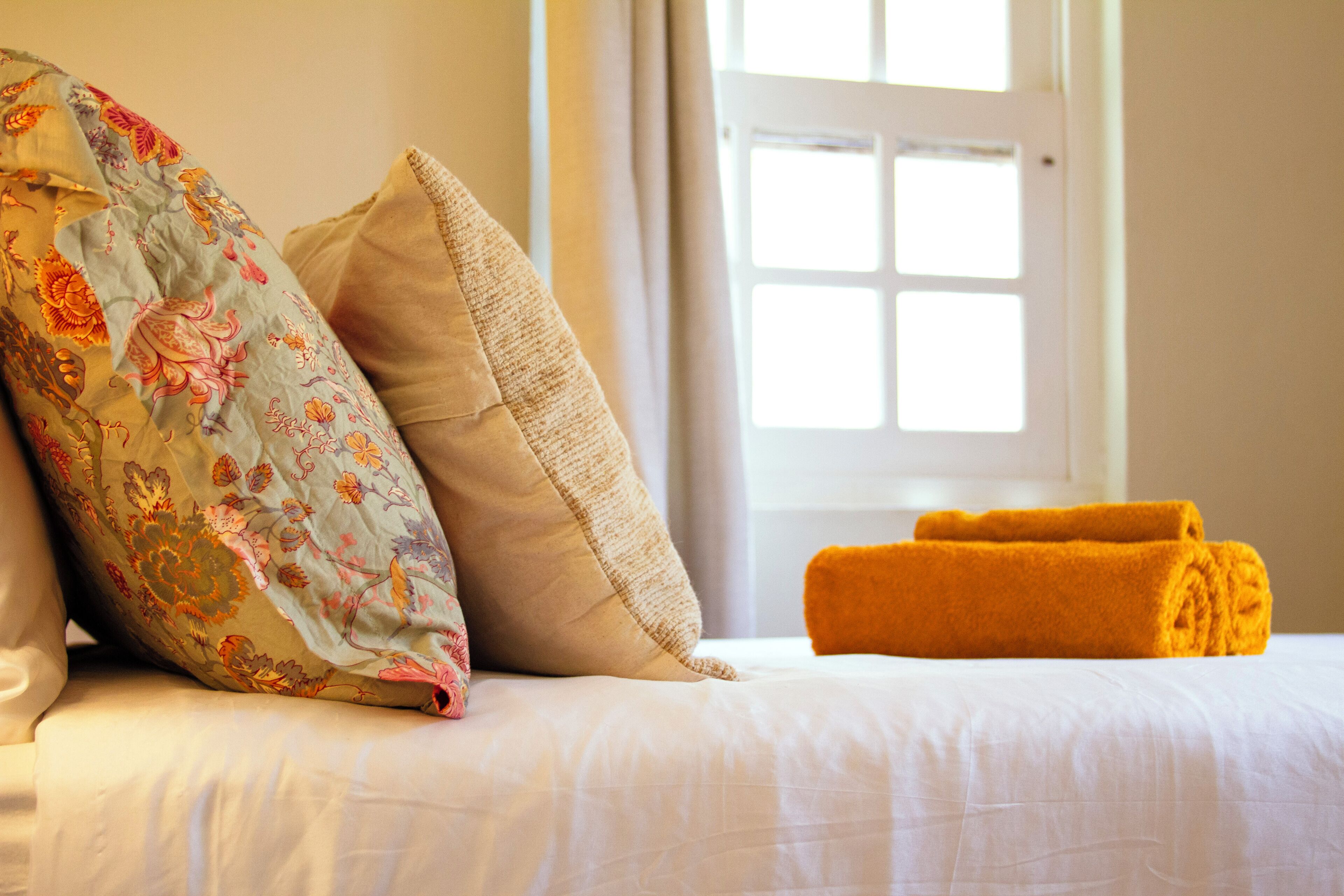 Ein Bett mit blumigen und beigen Kissen, ein aufgerolltes gelbes Handtuch und ein Fenster mit Blick.