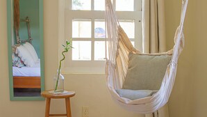 Una habitación con una silla hamaca, un taburete de madera con un jarrón y una cama con almohadas.