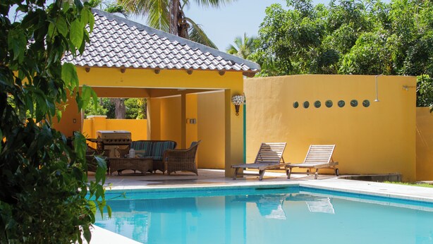 Zona de piscina con un edificio amarillo, sillas de descanso y una zona de estar cubierta.