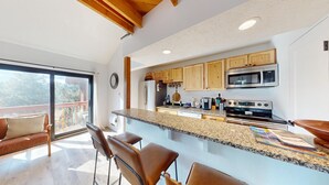 Una cocina moderna con encimeras de granito, armarios de madera y electrodomésticos de acero inoxidable. Se observa una isla grande con taburetes de bar y una puerta corrediza de vidrio que da a un balcón.