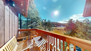 Una terraza de madera con un banco y una mesa, con vista a un área boscosa y un cielo despejado.