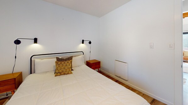 A bedroom with a bed, two bedside tables, a nightstand, a wall-mounted heater, and a door leading to another room.