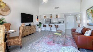 Een moderne woonkamer met een bank, een eethoek met rieten stoelen, een keuken met barstoelen en een plafondventilator.
