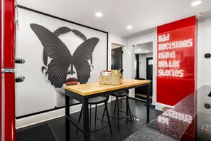 Ein moderner Arbeitsraum mit einer großen Wandmalerei, einem Holztisch mit zwei Stühlen und einer roten Akzentwand mit Neon-Schrift.