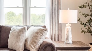 A brown leather sofa with two white throw pillows, a white lamp with a distressed design, and a view through a window with curtains.