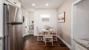 A kitchen with a stainless steel refrigerator, wooden cabinets, a dining table with chairs, and a 'gather here' sign on the wall.