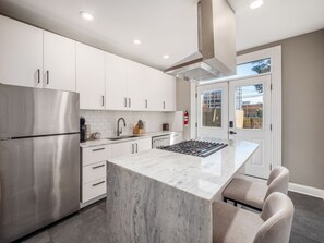 A modern kitchen with white cabinets, a stainless steel refrigerator, a marble countertop, and a gas range.