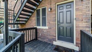 A brick building with a black door and window shutters, a wooden staircase, and a metal railing.