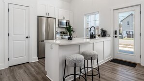 Una cocina moderna con armarios blancos, un refrigerador de acero inoxidable, un microondas y una cafetera. Hay dos taburetes en la isla y una ventana que proporciona luz natural.