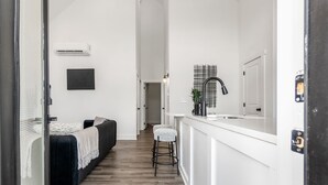 Un pasillo estrecho con una cama, una pequeña sala de estar, un mostrador blanco con un lavamanos y una obra de arte abstracta negra en la pared.