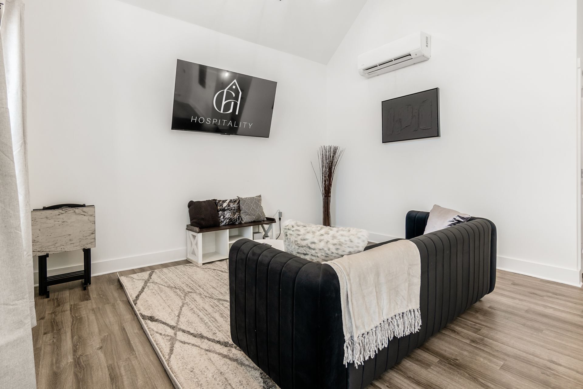 Un dormitorio moderno con un sofá negro, una mesita de noche blanca, un banco y un televisor grande montado en la pared que muestra el logotipo de un hotel.