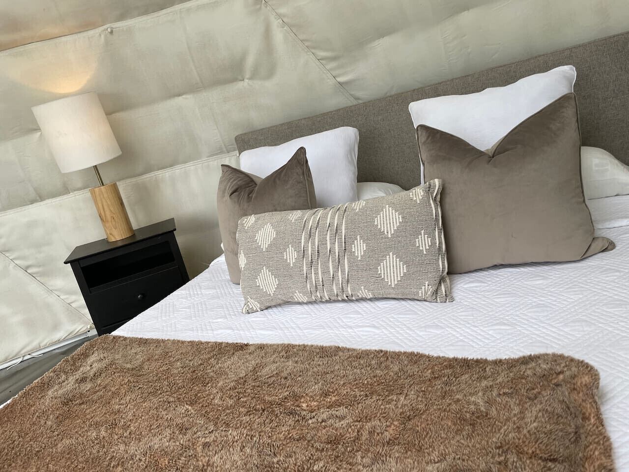 Una cama bien hecha con edredón blanco, una manta marrón y tres almohadas en tonos blancos y marrones.
