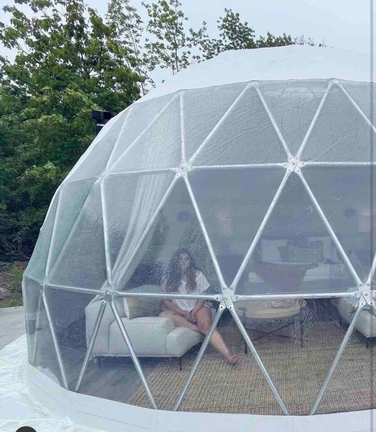 Una estructura de cúpula geodésica con cubierta transparente, un área de estar acolchada de color blanco en su interior y vistas a árboles en el fondo.