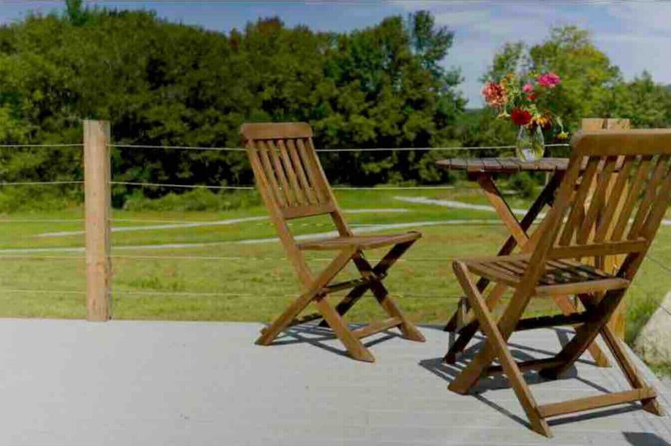 Two wooden chairs and a table on a deck with a view of a grassy field and trees.