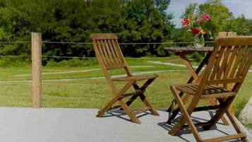 Two wooden chairs and a table on a deck with a view of a grassy field and trees.