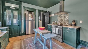 A kitchen with a green door labeled "PANTRY," stainless steel appliances, a wooden table with a red bowl, and a patterned backsplash.