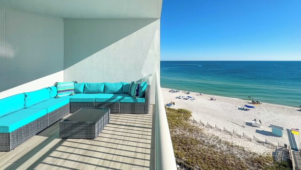 A balcony with a wicker sofa and a coffee table, overlooking a beach with umbrellas and a clear blue sky. - Palacio 703 | Oceanfront Resort Amenities | My Beach Getaways (Pensacola)