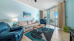 A living room with a sofa, armchair, coffee table, and a view through a sliding glass door to the outside. - Palacio 703 | Oceanfront Resort Amenities | My Beach Getaways (Pensacola)