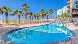 A swimming pool with lounge chairs and palm trees, surrounded by a white fence and a building with balconies. - Tidewater 801 | Pools, Gym, Hot Tub, Wifi | Private Balcony | Ocean Front Views (Orange Beach)