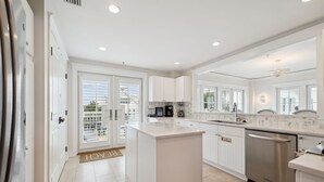 A modern kitchen with white cabinetry, a central island, stainless steel appliances, and a large window providing natural light. - Beach Front + Pool Front Watercolor Condo | Beach Views of 30A (Watercolor)