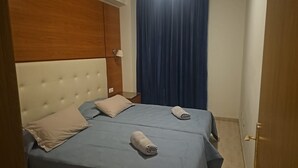 A double bed with two pillows and two rolled towels, a wooden headboard, a nightstand with a lamp, and a blue curtain.