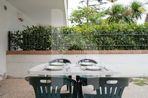 An outdoor dining area with a table set for two, surrounded by green chairs, against a white wall with a black metal fence and greenery in the background. - Prestige - Riva - Ground floor apartment with patio (Pineto)