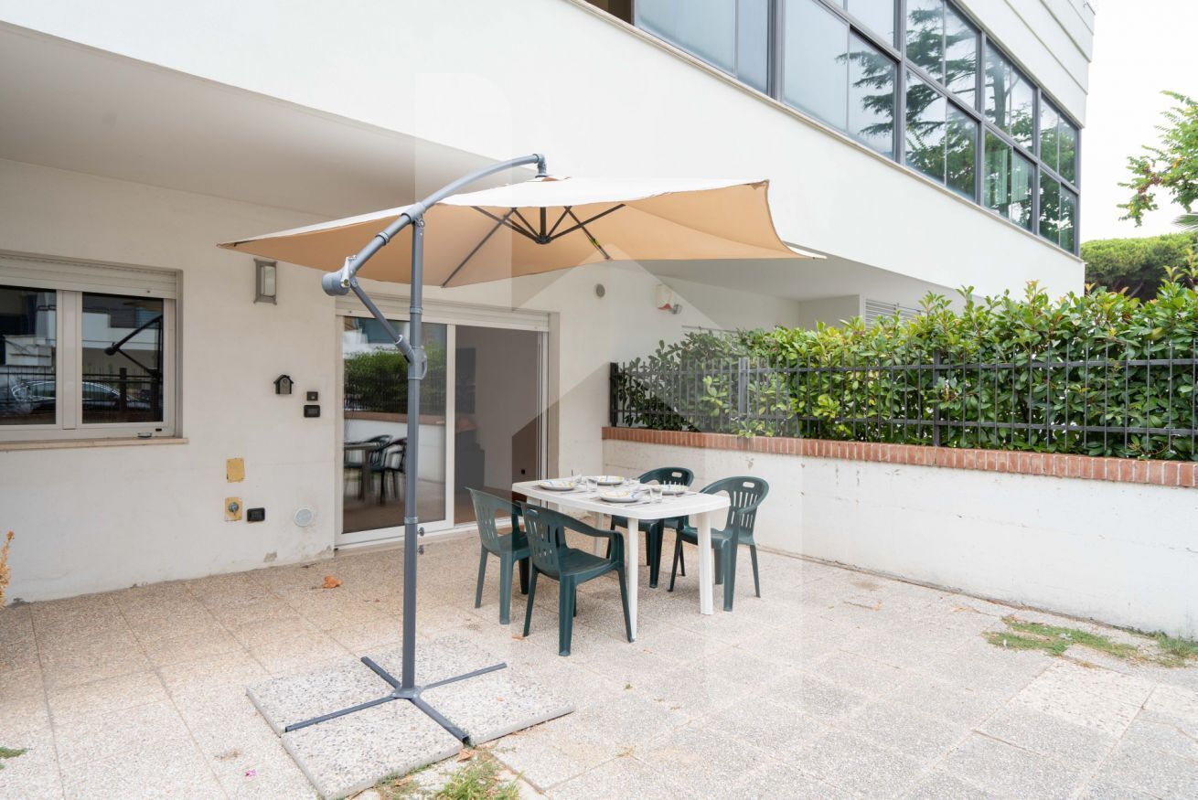 An outdoor patio with a table and chairs, a large umbrella, and a modern building in the background.