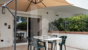 Un espace patio aménagé avec une table et des chaises, un grand parasol, et une vue sur un jardin et l’entrée d’un bâtiment.