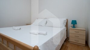 Un lit bien fait, avec du linge de lit blanc et des serviettes pliées, une tête de lit en bois, une petite table de chevet avec une lampe turquoise et une commode.