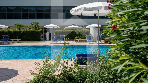 Une piscine entourée de chaises longues et de parasols, avec de la verdure tout autour et un bâtiment en arrière-plan.