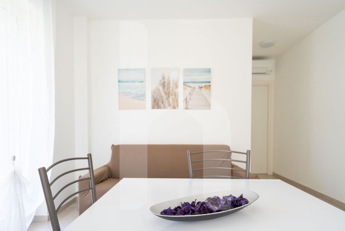 Zona pranzo con tavolo bianco, due sedie, un divano color beige e una parete con tre quadri incorniciati.