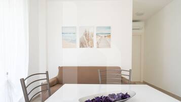Zona pranzo con tavolo bianco, due sedie, un divano color beige e una parete con tre quadri incorniciati.