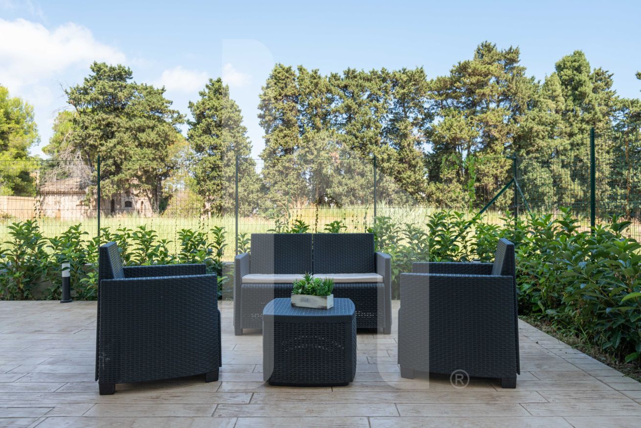 An outdoor seating area with wicker furniture and a small table, surrounded by greenery and trees.