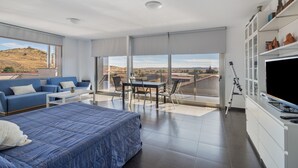 A modern hotel room with a bed, a sofa, a dining table, and a TV.