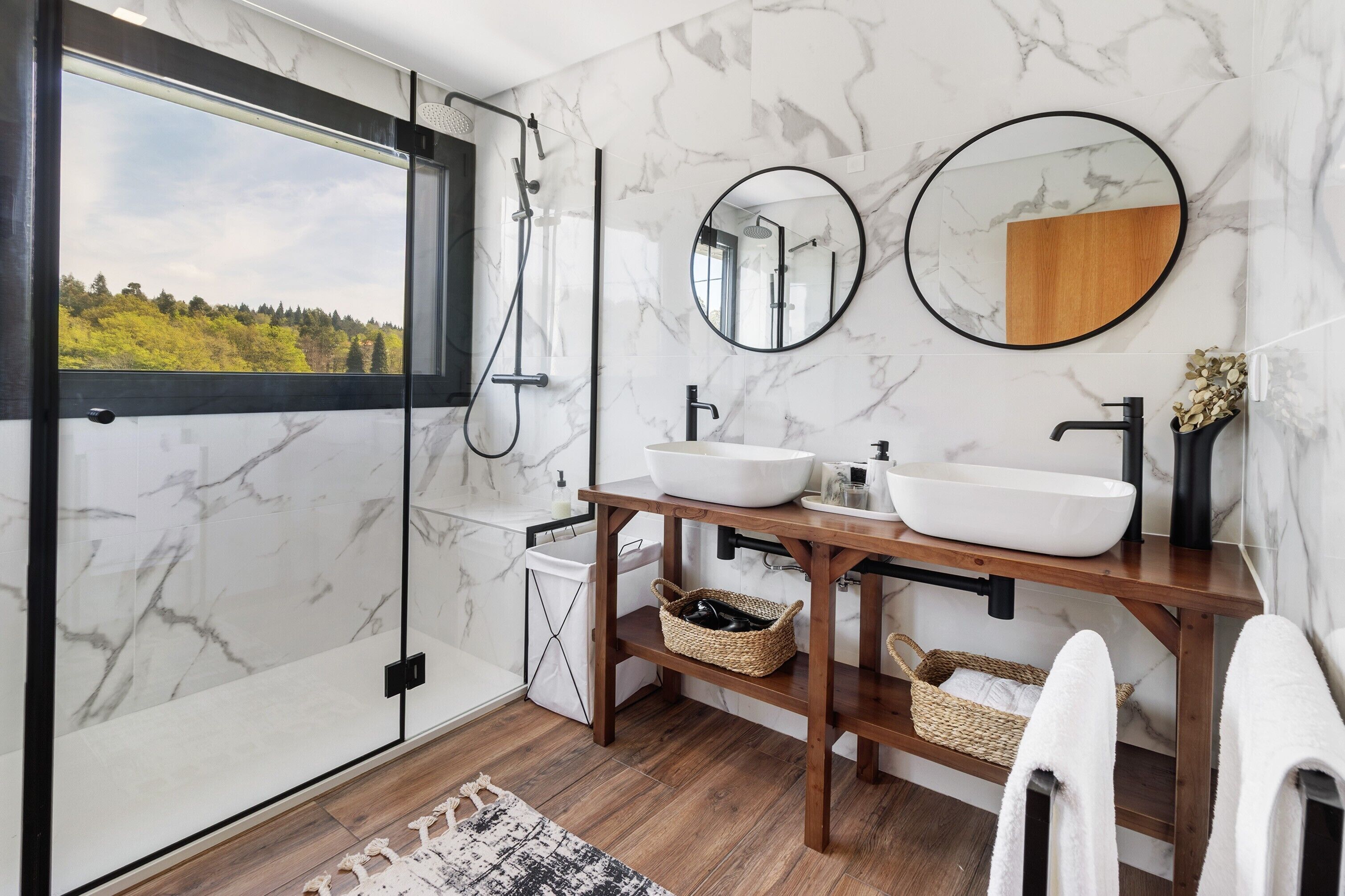 Ein Badezimmer mit großem Fenster, einem Holzwaschtisch, einem weißen Waschbecken, zwei Spiegeln und einer Dusche.