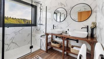 Ein Badezimmer mit großem Fenster, einem Holzwaschtisch, einem weißen Waschbecken, zwei Spiegeln und einer Dusche.