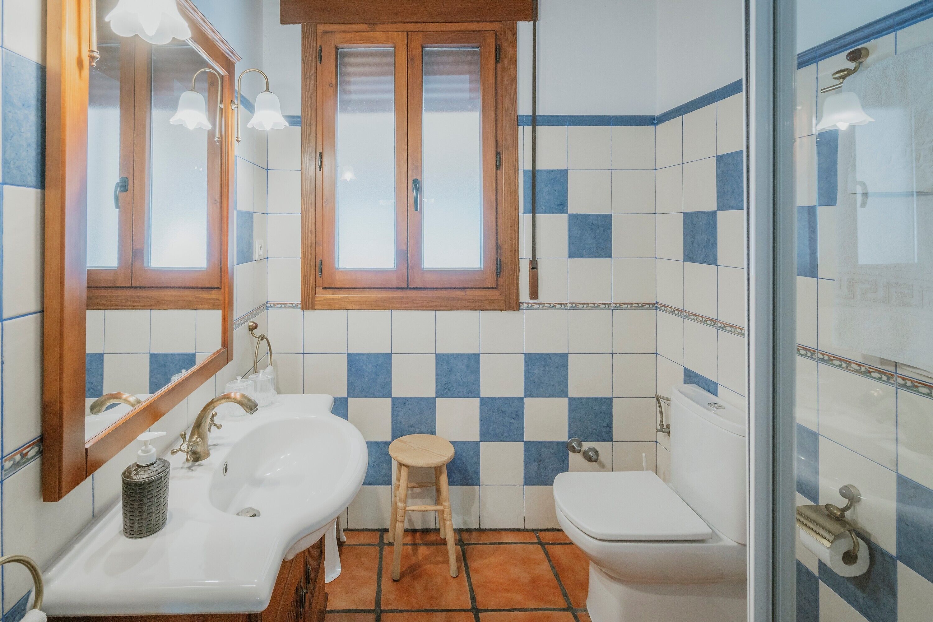Un baño con un lavamanos blanco, un inodoro, un taburete de madera y una ventana con marco de madera.