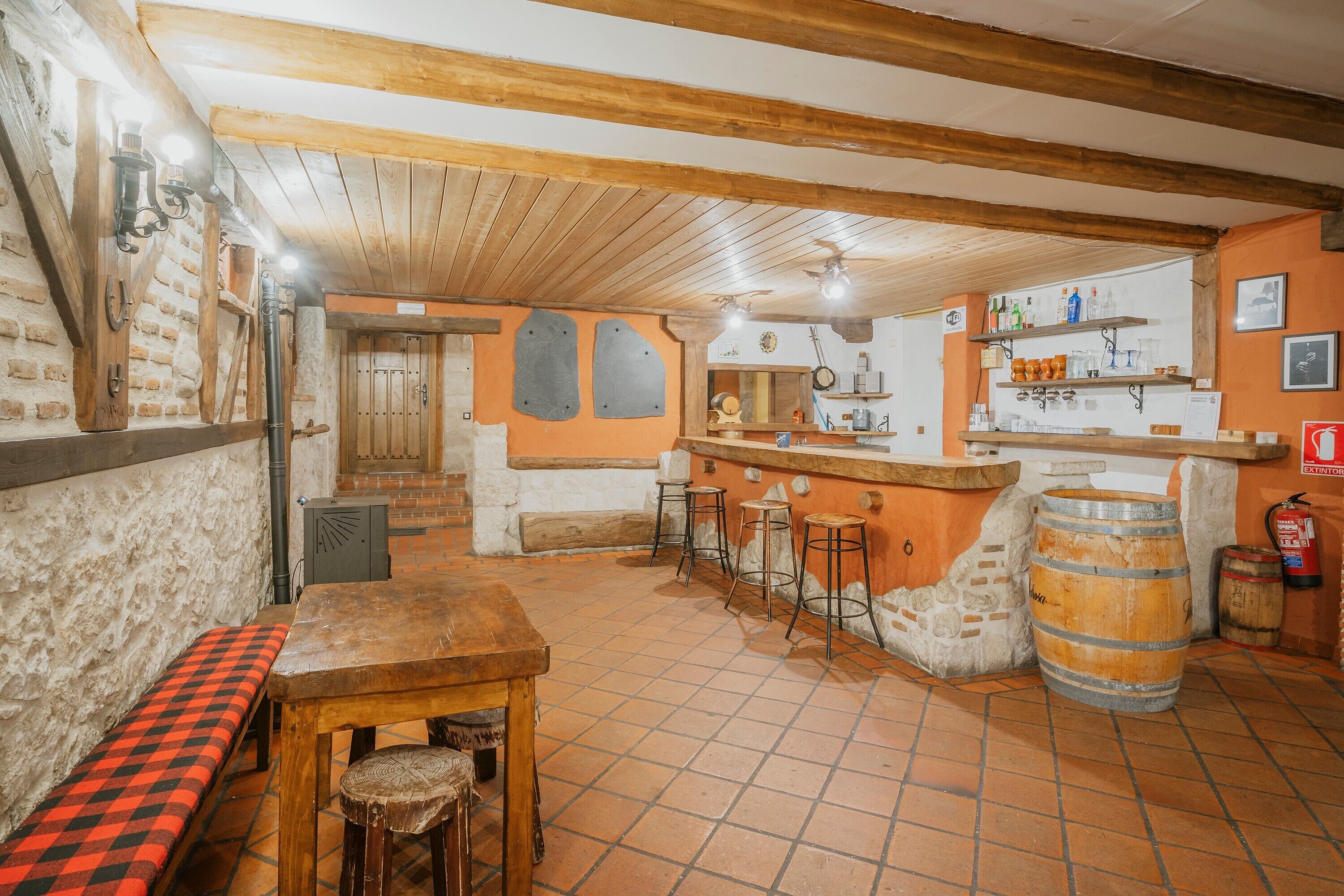 Un interior rústico con una mesa de madera, taburetes de bar, un banco, suelo de baldosas y una zona de bar con estantes y botellas.