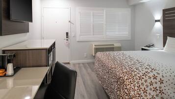 A hotel room with a bed, a desk, a chair, a TV mounted on the wall, a window with blinds, and a bedside table with a lamp.