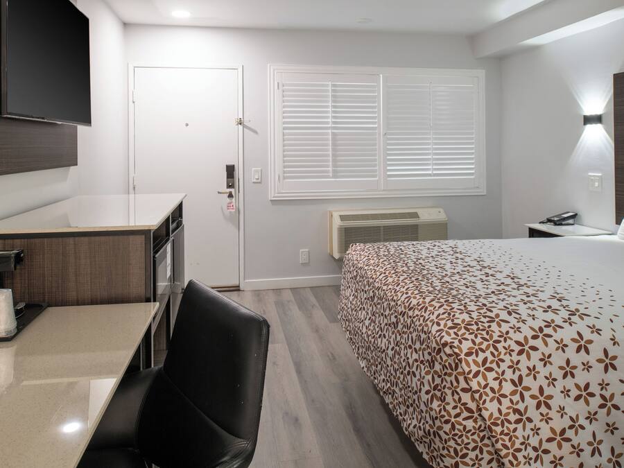 A hotel room with a bed, a desk, a chair, a TV mounted on the wall, a window with blinds, and a bedside table with a lamp.