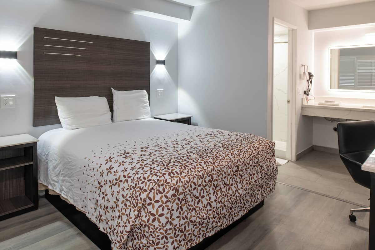 A modern hotel room with a large bed, bedside tables, a desk with a chair, and a bathroom visible through an open door.
