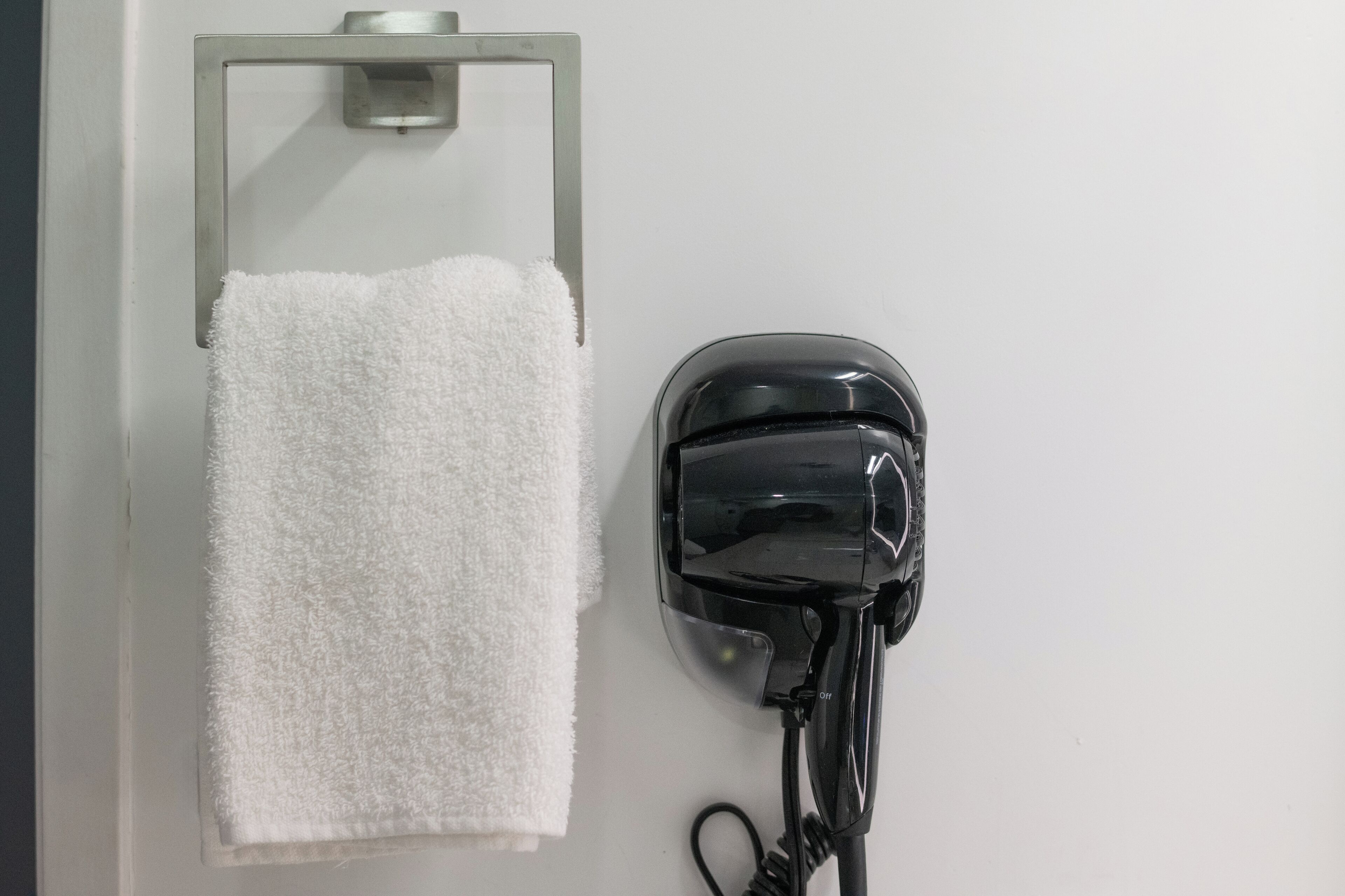 A white towel hanging on a metal towel rack next to a black hairdryer on a white wall.