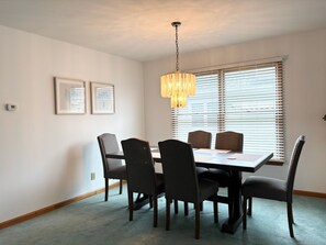 A dining room with a rectangular table, six chairs, a hanging light fixture, and two framed pictures on the wall.