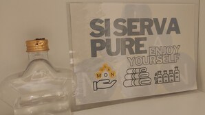 A clear glass bottle with a gold cap, next to a framed sign with the text "SISERVA PURE ENJOY YOURSELF" and a hand holding a bottle. - Bèz Apartment in Milan Tortona street Fashion design district  (Milano)